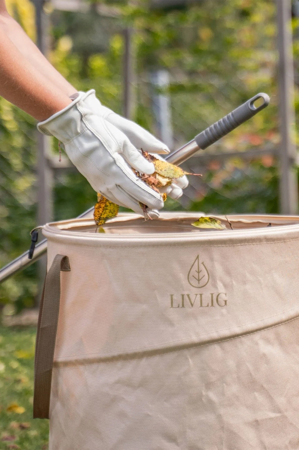 Eine Person mit Gartenhandschuhen wirft Laub und Pflanzenschnitt in den geöffneten beigen LIVLIG Pop-up Gartensack. Der 90-Liter Gartenabfallsack aus robustem Oxford-Gewebe steht stabil auf dem Rasen.