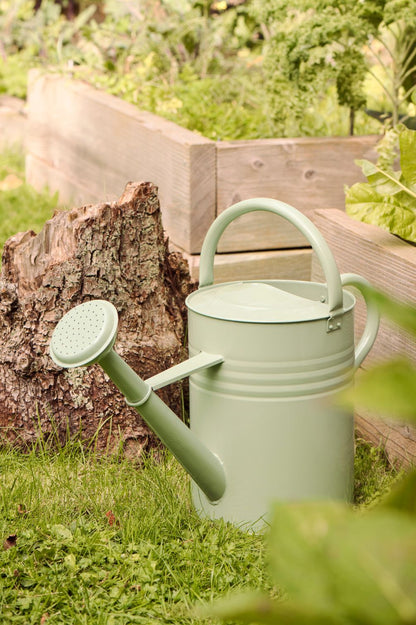 Pastellgrüne Giesskanne aus Metall im Gras vor Hochbeet und Baumstamm im naturnahen Garten.
