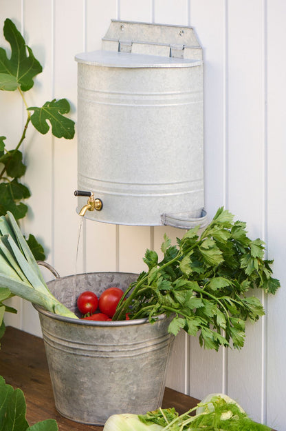 Wand-Wasserbehälter aus Metall mit Messinghahn über Zinkwanne mit Tomaten und Kräutern.