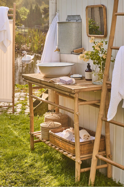 Wand-Wasserbehälter mit Hahn aus Metall in pastellgrün über Emaille-Schüssel auf Bambustisch im Garten.