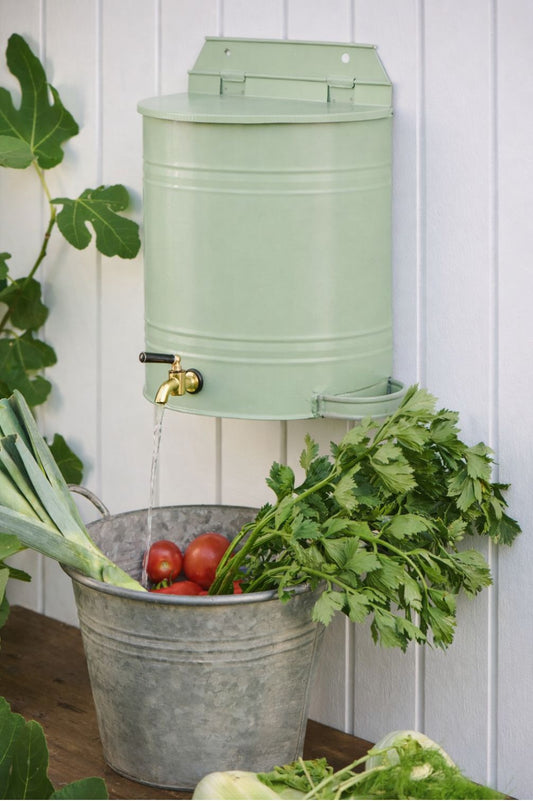Wand-Wasserbehälter aus Metall in pastellgruen mit Messinghahn über Zinkwanne mit Tomaten und Kräutern.