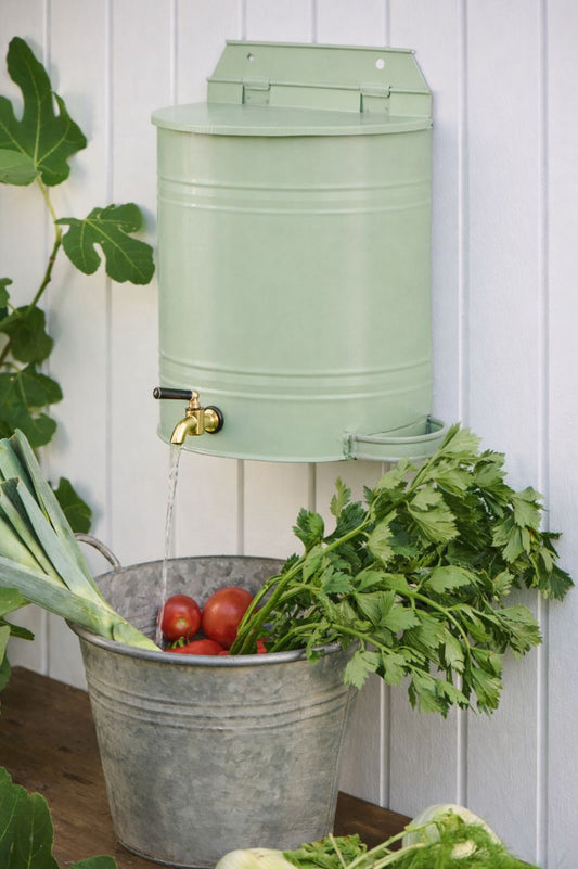 Wand-Wasserbehälter aus Metall in pastellgruen mit Messinghahn über Zinkwanne mit Tomaten und Kräutern.